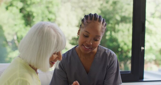 African American caregiver in scrubs showing senior hand exercise on sofa by window after pause