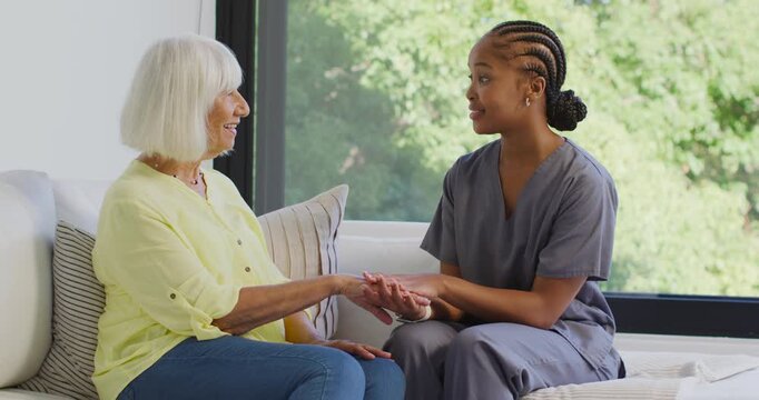 African-American nurse in scrubs turning holding Senior woman's hands soothing on couch by window