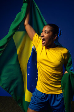 Adult woman in yellow jersey, blue shorts cheering, wrapping Brazil flag in studio lights on turf