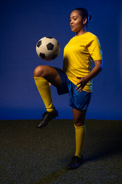 Woman balancing on one leg controlling soccer ball near knee in studio turf wearing yellow jersey
