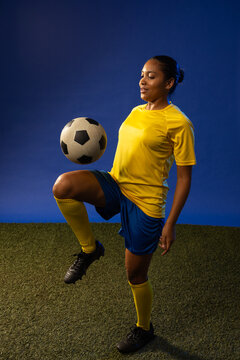 Adult woman balancing soccer ball on raised knee, posing on studio turf in yellow jersey