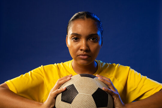African American woman posing against deep-blue backdrop wearing yellow jersey holding soccer ball