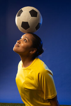 African American woman balancing soccer ball on forehead in studio with yellow jersey and turf