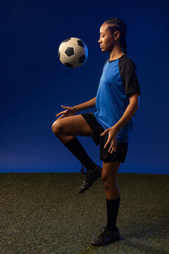 African female athlete balancing classic paneled soccer ball near raised knee on turf in blue kit