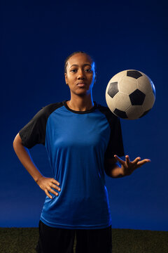 African American female athlete balancing soccer ball with right hand on studio turf in blue kit