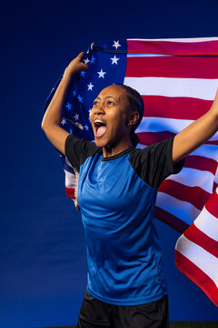Female athlete standing in studio holding United States flag while wearing blue jersey and shorts