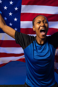 African American female athlete wearing blue jersey, raising arms, cheering before US flag on turf