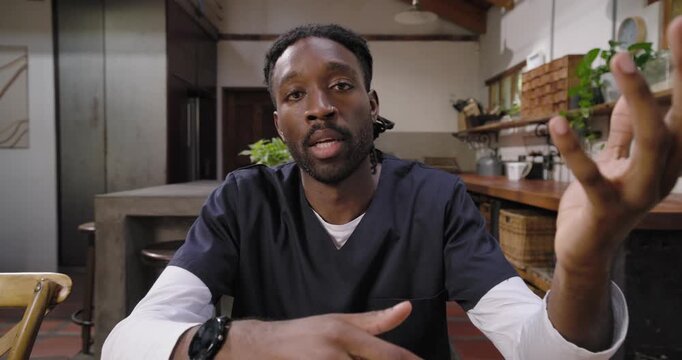 Adult African man in navy scrubs answering camera at kitchen table on health leaning checking hands