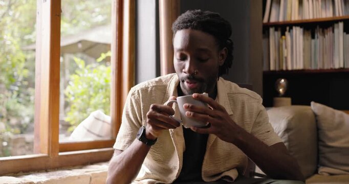 Adult African man sitting on sofa by wooden window with mug light-drawing gaze sipping smiling cam