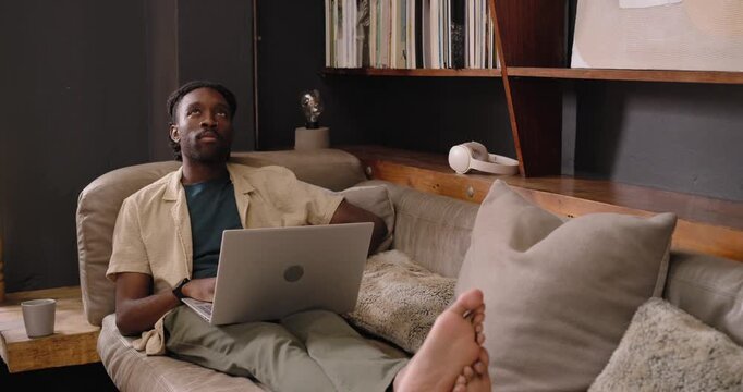 Adult African man reclining on sofa at home finishing laptop work closing laptop stretching resting