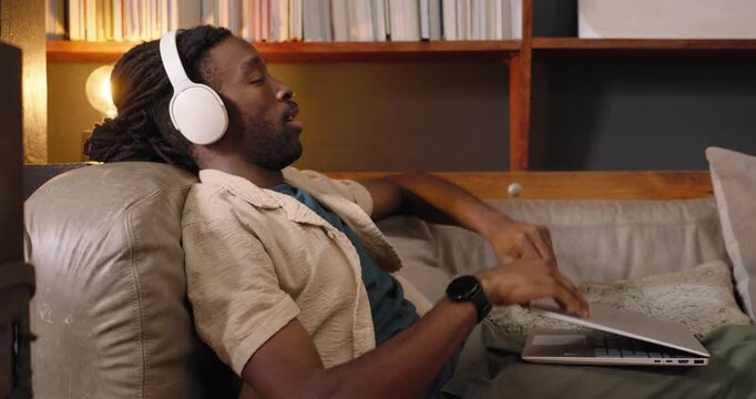 African man with headphones finishing laptop work, closing lid and leaning back on couch to relax