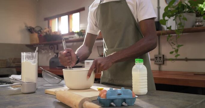 African American man in olive apron whisking batter at kitchen island after assembling eggs milk