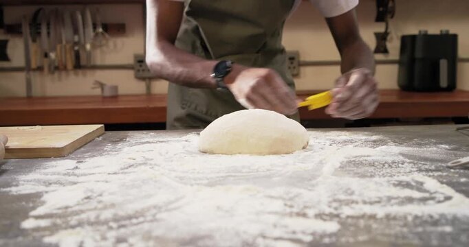 Male baker in apron with floured dough shaping, brushing on counter using yellow scraper for baking