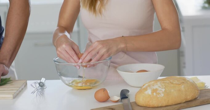 Couple in bright kitchen, adult woman tapping and cracking eggs into clear glass bowl making batter