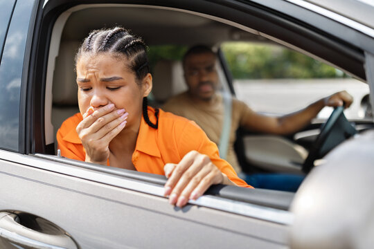 Young Black woman feeling carsick leaning out open car window, concerned man driving in background, motion sickness and nausea during road trip lifestyle concept