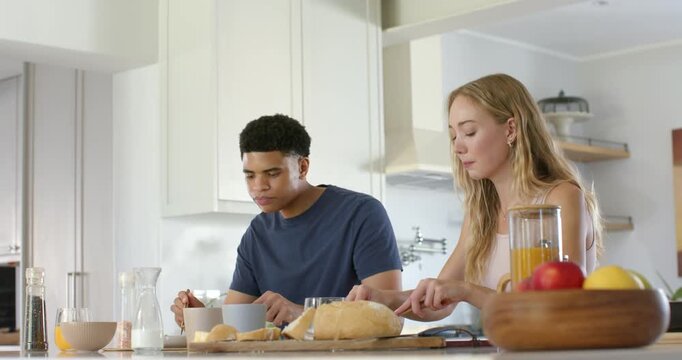 Diverse couple at counter, woman taking knife slicing bread as man stirring bowl making breakfast