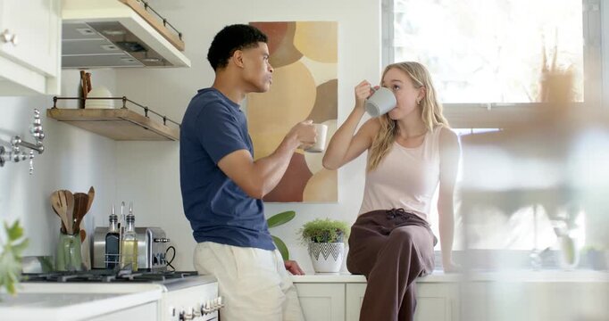 Couple leaning on kitchen counter with man initiating chat as woman sitting and sipping from mugs