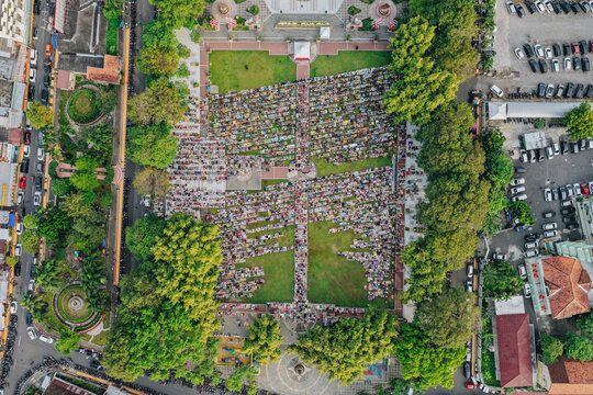 Aerial view of a large crowd of people gathered for communal prayer in a green city square surrounded by lush trees and urban streets in Klaten, Jawa Tengah, Indonesia.