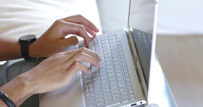 Man typing on laptop on home sofa after hands poised, wearing wristwatch, using touchpad for work