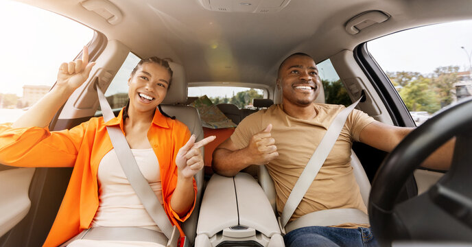 Cheerful young African American couple listening music and dancing inside car, expressing happiness, freedom and fun during road trip or daily commute