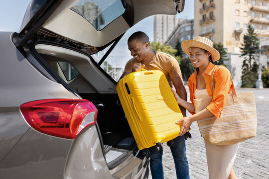 Happy young Black couple loading yellow suitcase into car trunk, woman in straw hat smiling beside man lifting luggage, travel departure and vacation lifestyle concept
