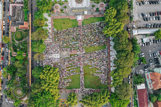 Aerial view of a large crowd of people gathered for mass prayer in a public square surrounded by lush green trees and parking lots in Klaten, Jawa Tengah, Indonesia.