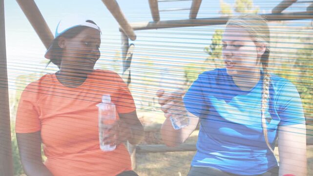 Two women arriving on wooden bench under bars, smiling, reaching bottles, hydrating for fitness