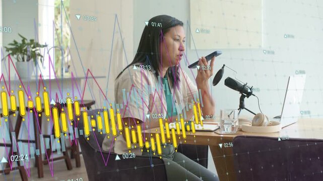 Podcast host speaking into phone and mic, driving finance graphs scrolling over laptop and notebook