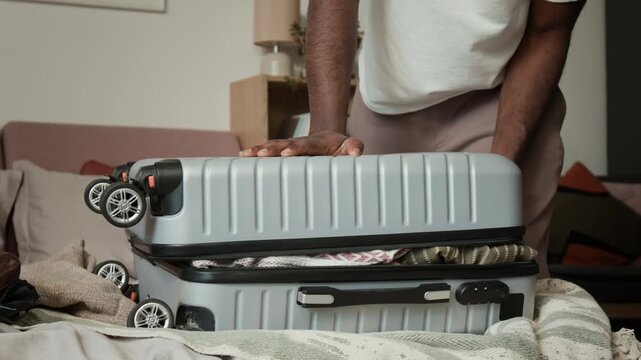 Midsection shot of unrecognizable black man struggling to zip up stuffed suitcase for vacation or overseas journey, pressing down with knee and body weight to comply with luggage restrictions