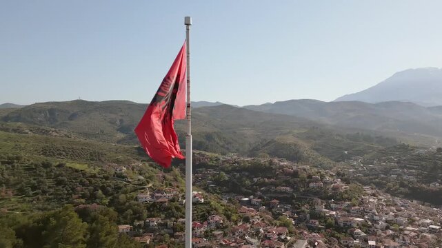 Aerial view Albanian national flag prominently wave atop stone fortification at Berat Castle - Kalaja e Beratit under clear blue sky, overlooking lush, forested hillside and Berat cityscape