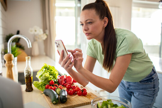 Young woman checking recipe on smartphone while preparing salad in home kitchen