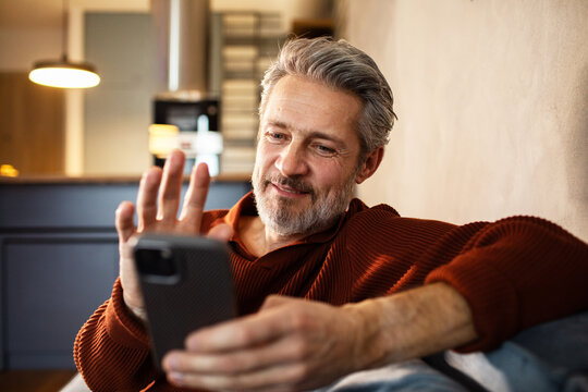 Smiling middle-aged man waving on smartphone video call at home