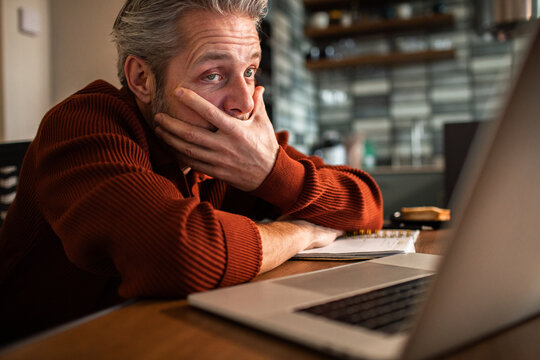 Middle-aged man stressed while working on laptop in home kitchen