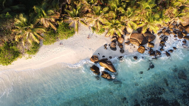 Top-down drone seascape showing turquoise water, sandy shore, and boulders along a lush coastline.
