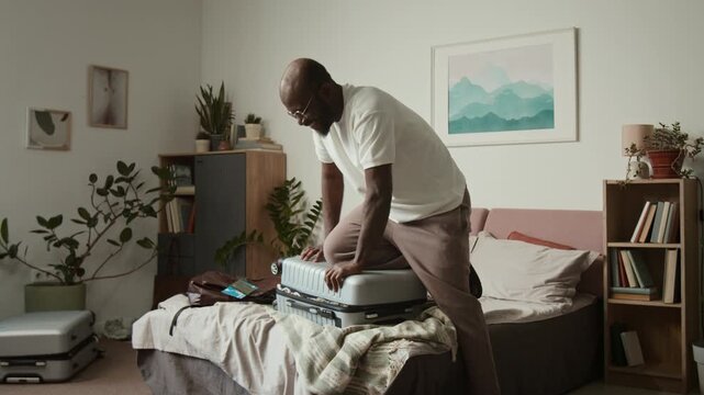 Full shot of cheerful African American man closing suitcase stuffed full with clothes and personal belongings for traveling on holiday, pressing with foot, sitting on lid and struggling to zip luggage