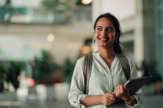 Indian female student smiling confidently, moving through a modern university hall, carrying a laptop and backpack