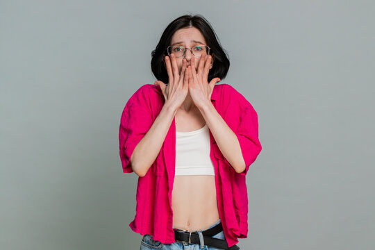 Young woman shivers hugs her shoulders and anxiously looks around, clearly expressing nervousness or fear. Girl demonstrates intense worry and apprehensive emotional state, isolated on gray background