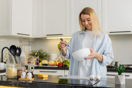 Middle-aged woman whisking eggs in bowl in kitchen making omelet batter for healthy fit meal. Young girl learning easy simple home cooking from recipe video for clean eating diet meal prep nutrition.