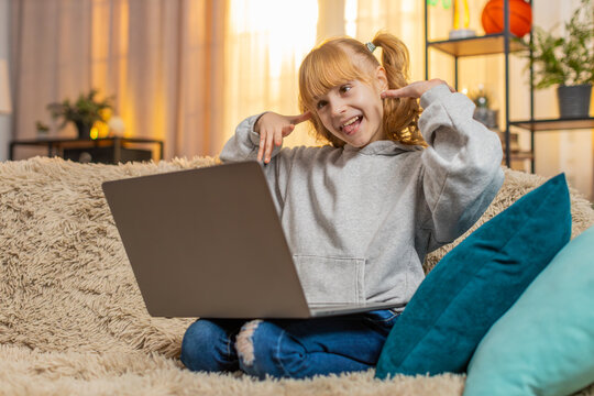 Young teenager girl rests at home with laptop, making a fun video call online and laughing at her own reflections making silly faces. Child kid waves hi in excited joy and saying I am so funny look
