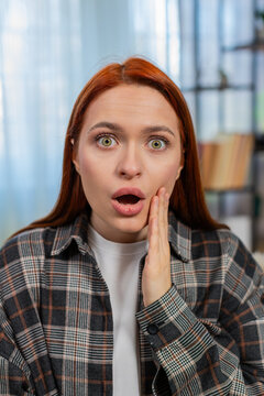 Young woman at home raising eyebrows and opening mouth with surprise after seeing something unexpected. Caucasian redhead girl on sofa reacting emotionally, shocked and amazed by sudden information