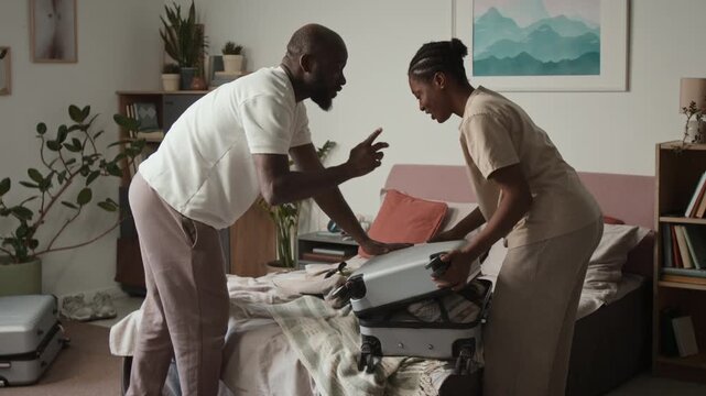 Medium full shot of African American couple packing belongings into suitcase, adding scuba diving mask, husband pushing down while wife zipping up lid, for traveling abroad for vacation