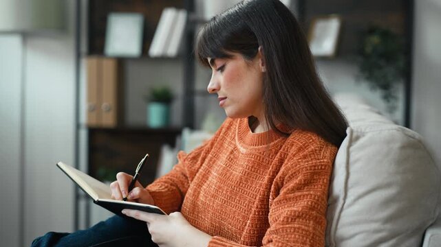 Video of beautiful writer taking notes in a notebook while sitting on the couch at home