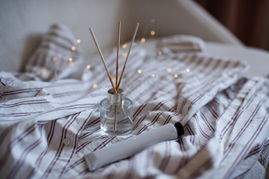 Home aroma liquid glass bottle diffuser with sticks on pajama shirt in bed with glowing lights. Hygge cozy atmosphere.