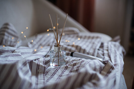 Home aroma liquid glass bottle diffuser with sticks on pajama shirt in bed with glowing lights. Hygge cozy atmosphere.