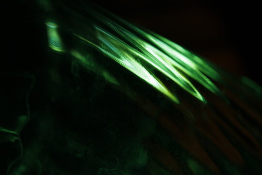 Primer plano abstracto de un vaso de vidrio con bordes labrados, iluminado con luz verde intensa sobre fondo oscuro. Las facetas del cristal generan reflejos c&aacute;lidos y l&iacute;neas luminosas brillantes.