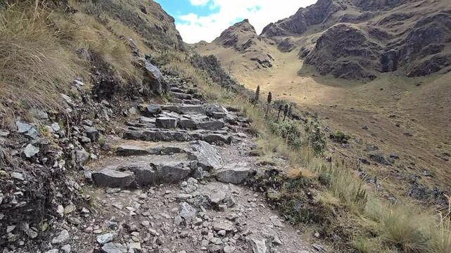 Dead Woman&rsquo;s Pass, Peru - 26 June 2024. First-person POV walking along stone steps on the Inca Trail, with rocky terrain, dry grass, and steep mountain slopes under a partly cloudy sky.