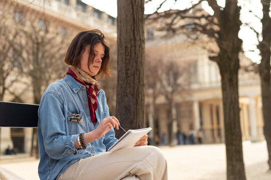 A woman sits on a bench in a city square drawing in a sketchbook. She is wearing a denim shirt and a red scarf with buildings in the blurred background.