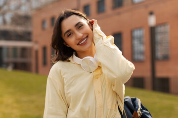 Fototapeta premium Pretty happy lady student posing outdoors at university campus, wearing casual outfit, wireless headphones and bag, smiling at camera