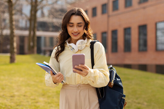 Cheerful lady student with smartphone and workbooks standing outdoors in college campus after classes, chatting with friends and smiling