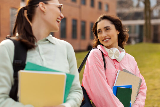 College friendship. Two happy female students chatting outdoors after classes, holding workbooks and smiling to each other, walking on campus area